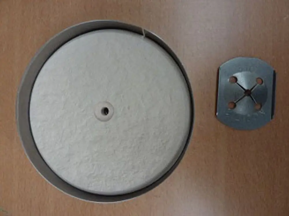Reparatursatz von Remeha mit einer runden, hellen Fläche in einer Metallfassung und einem metallischen Montageelement mit vier Öffnungen daneben.